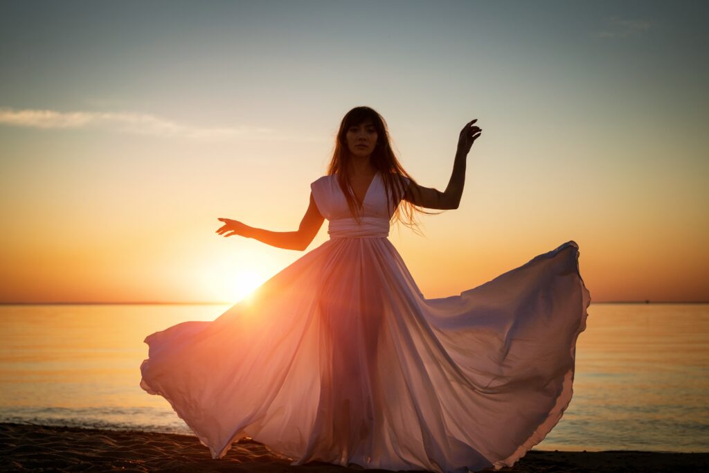 Beautiful female silhouette on the seashore in a white long dress at sunset